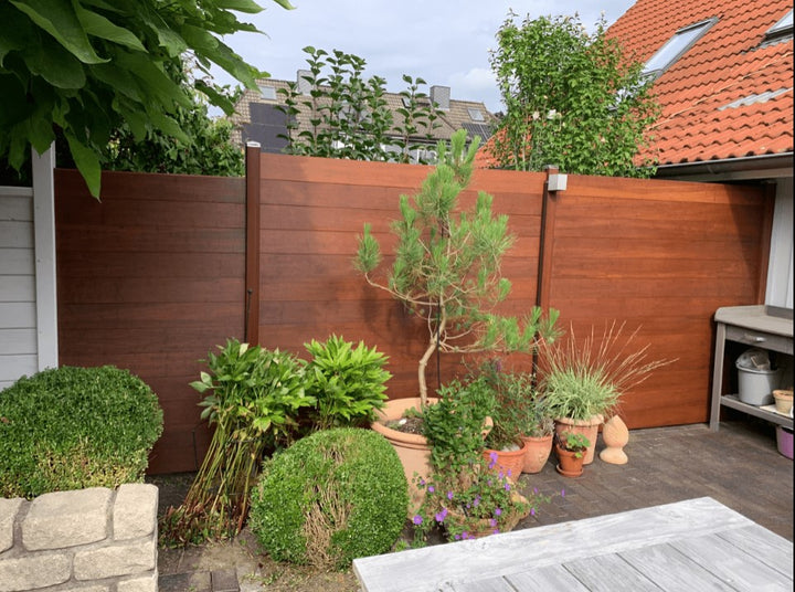 Moderner AMBOO Bambus-Sichtschutzzaun in warmer Optik mit dunklen Pfosten, der eine Terrasse begrenzt und als Hintergrund für diverse Gartenpflanzen und einen Pflanztisch dient.