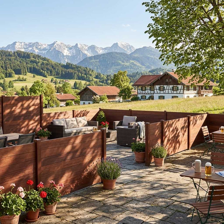 Terrasse mit coffee Holz-Sichtschutzwänden, Sitzmöbeln und Topfpflanzen; dahinter liegen Wiesen, Bauernhäuser und ein Alpenpanorama.