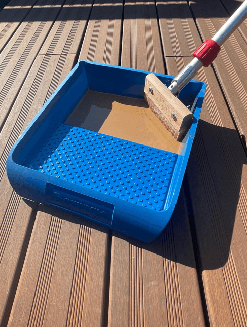 Blaue Farbwanne mit Holzflächenstreicher und Pflegeöl auf einer braunen Holz-Terrasse. Das Streichwerkzeug ist an einem Verlängerungsstiel befestigt und eignet sich für das gleichmäßige Auftragen von Ölen und Pflegeprodukten auf Terrassenbelägen.