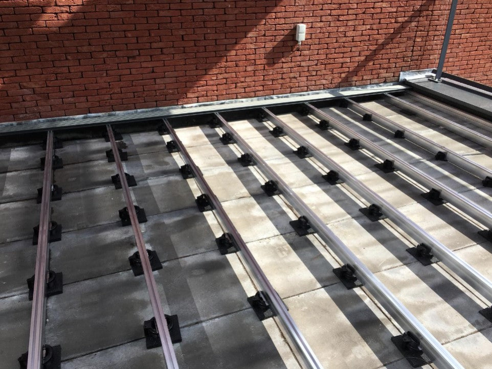 Terrassen-Unterkonstruktion aus Aluminium auf Stelzlagern vor einer roten Klinkerwand. Die Profile sind auf Betonplatten ausgerichtet und für die Verlegung von Terrassendielen vorbereitet.