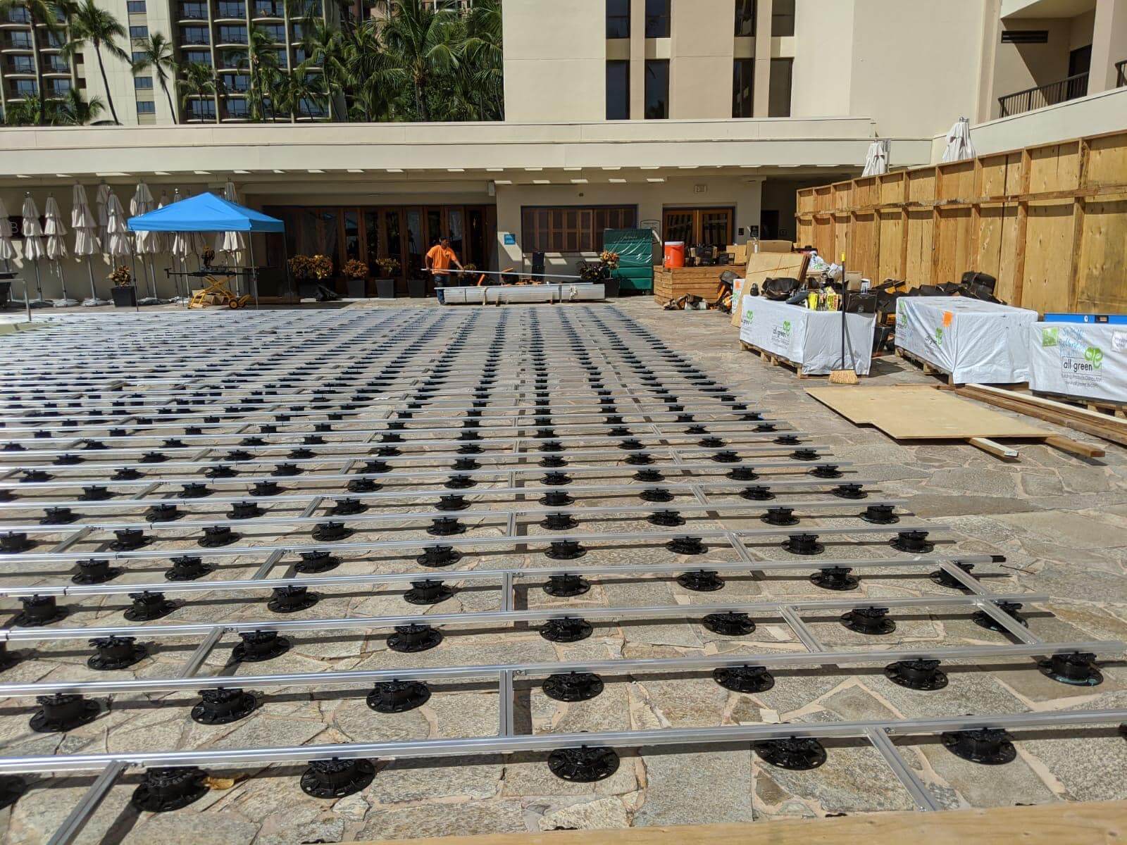 Terrassen-Unterkonstruktion aus Aluminium auf Stelzlagern vor einer roten Klinkerwand. Die Profile sind auf Betonplatten ausgerichtet und für die Verlegung von Terrassendielen vorbereitet.