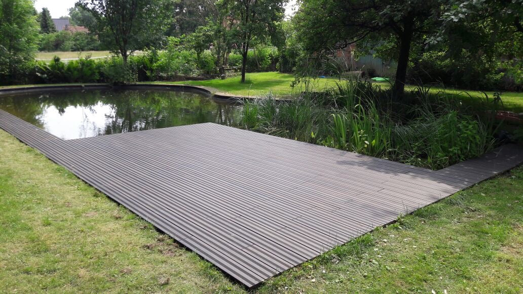 Grossige Bambus Terrasse mit schwarzen Terrassendielen direkt am Gartenteich eingebettet in eine grüne Gartenlandschaft mit Bäumen, Rasen und Uferbefpflanzung.
