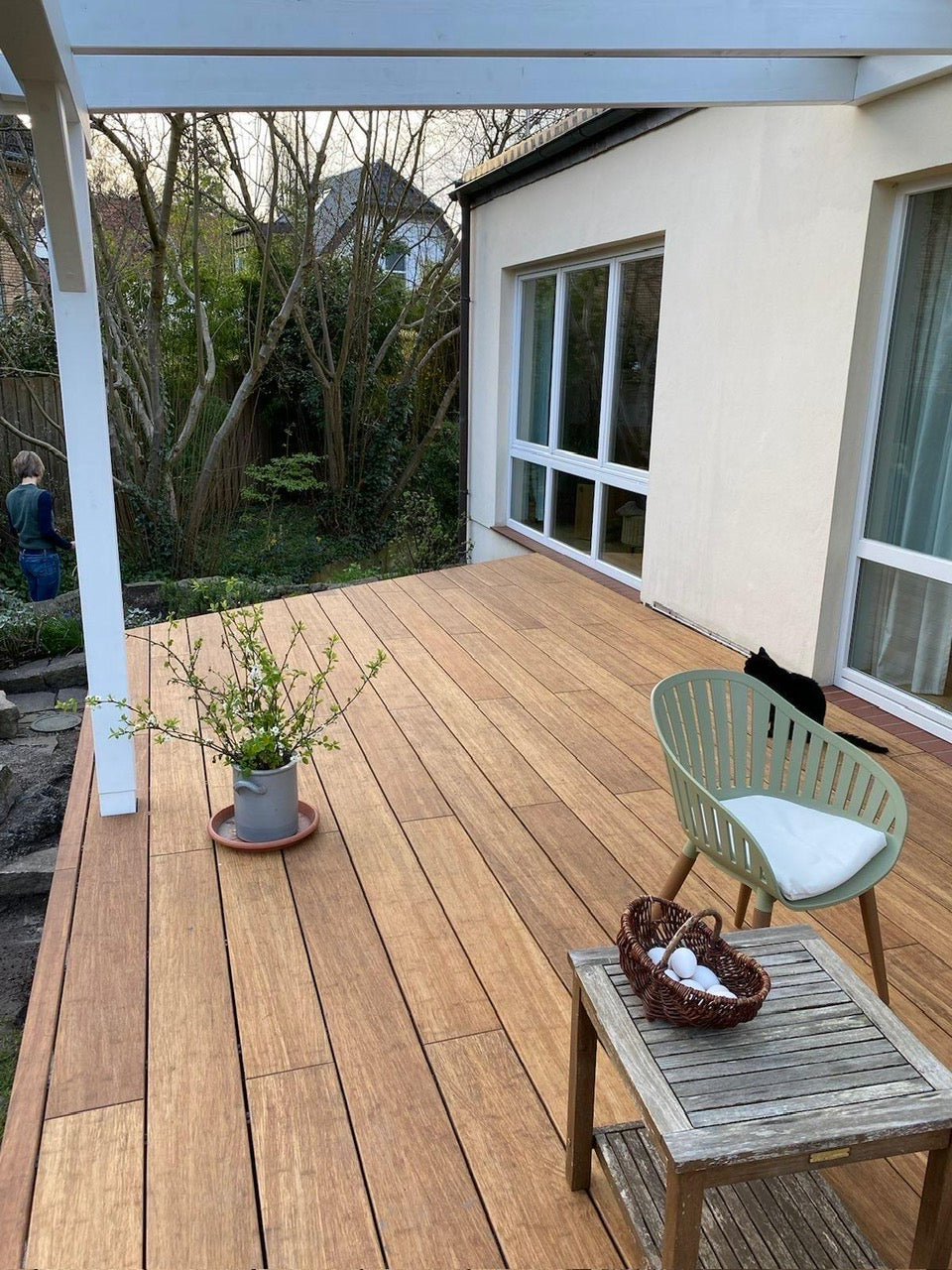 Moderne Terrasse mit hochwertigen glatten XL Bambus Terrassendielen, in der Farbe Sand  unter einer weißen Pergola. Gemütlicher Sitzbereich mit Gartenblick direkt am Wohnhaus mit großer Fensterfront.