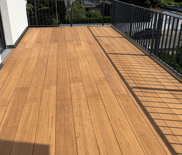 Großzügige Balkonterrasse, erstellt mit den AMBOOO Closed-Decking Dielen. Die Terrassendielen sind längs verlegt und werden von einem modernen anthrazitfarbenen Metallgeländer eingefasst. Sonnenschein erzeugt markante Schattenlinien des Geländers auf dem Terrassenboden. Im Hintergrund sind Wohnhäuser und Grünflächen zu sehen.