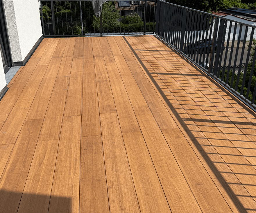 Großzügige Balkonterrasse, erstellt mit den AMBOOO Closed Decking Dielen. Die Terrassendielen sind längs verlegt und werden von einem modernen anthrazitfarbenen Metallgeländer eingefasst. Sonnenschein erzeugt markante Schattenlinien des Geländers auf dem Terrassenboden. Im Hintergrund sind Wohnhäuser und Grünflächen zu sehen.
