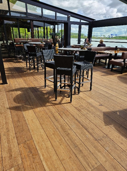 Überdachte Restaurantterrasse mit holzoptischem Terrassenbelag aus Closed Decking Dielen in warmen Sandtönen. Auf der Terrasse stehen zahlreiche Tische und schwarze Flechtstühle. Große Glasfronten und offene Seiten bieten einen weiten Blick auf die Umgebung. Das Sonnenlicht betont die natürliche Holzstruktur des Bodenbelags und schafft eine einladende Atmosphäre.