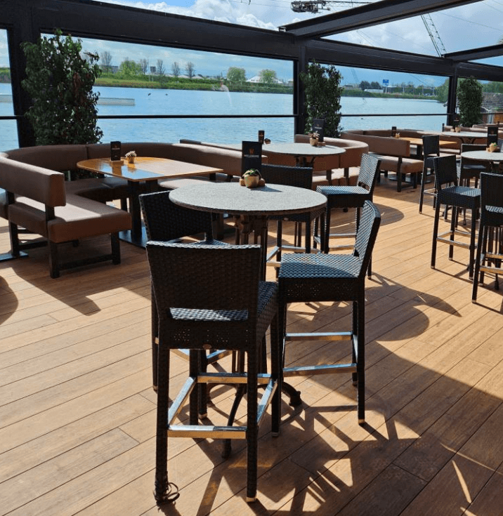 Überdachte Restaurantterrasse mit holzoptischem Terrassenbelag aus Closed Decking Dielen in warmen Sandtönen. Auf der Terrasse stehen zahlreiche Tische und schwarze Flechtstühle. Große Glasfronten und offene Seiten bieten einen weiten Blick auf die Umgebung. Das Sonnenlicht betont die natürliche Holzstruktur des Bodenbelags und schafft eine einladende Atmosphäre.