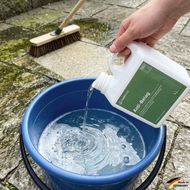 Eine Hand gießt AMBOOO Anti-Belag-Reiniger in einen mit Wasser gefüllten Eimer. Im Hintergrund sind eine Terrasse und ein Schrubber für die Reinigung von Außenflächen zu sehen.