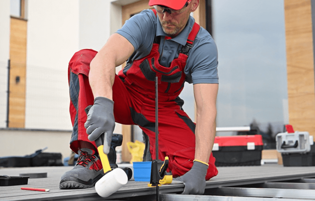 Handwerker in roter Arbeitskleidung montiert Bambus Terrassendielen auf einer Aluminium Unterkonstruktion mit einem weißen Gummihammer; im Hintergrund Werkzeugkoffer und modernes Gebäude. Die Montage der Bambus Terrassendielen auf einer Aluminium Unterkonsturktion.