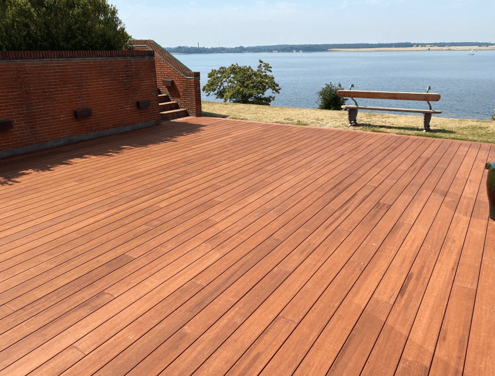 Große Terrasse aus rötlich-braunen Terrassendielen vor einer Backsteinmauer. Die Dielen verlaufen parallel über die gesamte Fläche und führen optisch zum angrenzenden See. Im Hintergrund befinden sich eine Sitzbank, eine Uferwiese und die ruhige Wasserlandschaft. Die Terrasse bietet viel Platz und verbindet hochwertige Holzoptik mit einer attraktiven Aussicht auf die Natur.
