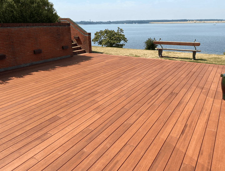 Große Terrasse aus rötlich-braunen Terrassendielen vor einer Backsteinmauer. Die Dielen verlaufen parallel über die gesamte Fläche und führen optisch zum angrenzenden See. Im Hintergrund befinden sich eine Sitzbank, eine Uferwiese und die ruhige Wasserlandschaft. Die Terrasse bietet viel Platz und verbindet hochwertige Holzoptik mit einer attraktiven Aussicht auf die Natur.