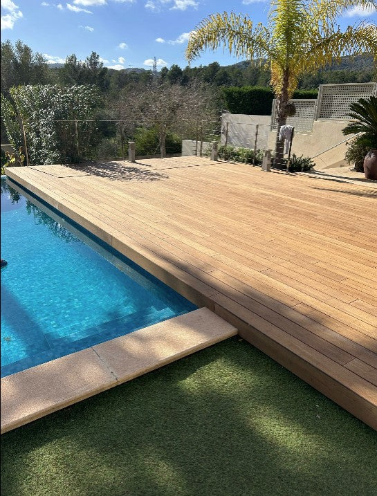 Eine weitläufige Terrasse aus hellen Sand Holzbambusdielen erstreckt sich entlang eines Swimmingpools mit klarem, blauem Wasser. Die Terrasse wird von Palmen, Sträuchern und Bäumen umgeben und schafft eine moderne, mediterrane Atmosphäre im Garten.