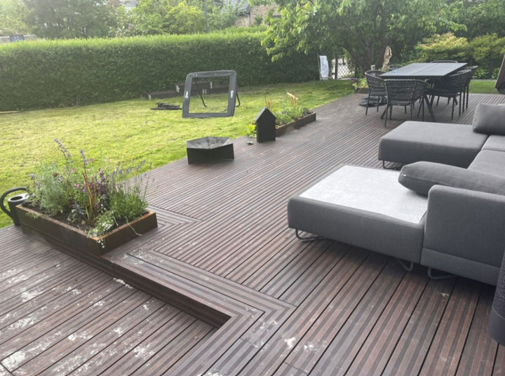 Eine großzügige AMBOOO Terrasse mit dunkelbraunen Bambus Terrassendielen erstreckt sich entlang eines gepflegten Gartens. Auf der Terrasse befinden sich eine moderne Lounge mit grauen Polstermöbeln, ein Esstisch mit mehreren Stühlen sowie bepflanzte Hochbeete. Die Terrasse geht nahtlos in eine weitläufige Rasenfläche über, die von Hecken und Bäumen umgeben ist und eine ruhige Gartenatmosphäre schafft.