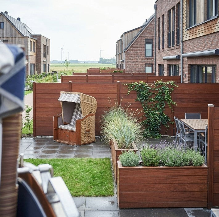 Terrasse zwischen modernen Wohnhäusern mit braunen Holz-Sichtschutzwänden, Strandkorb, Esstisch und bepflanzten Hochbeeten; im Hintergrund sind Felder und Windräder zu sehen.