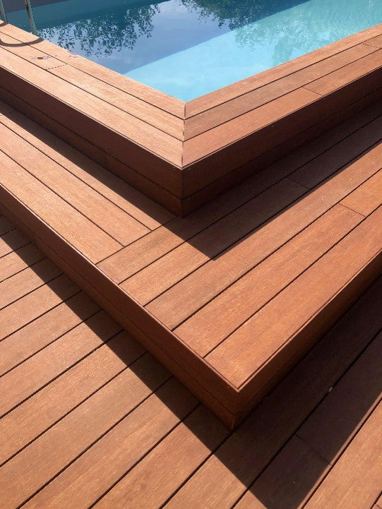 Detailaufnahme einer mehrstufigen Terrasse aus braunen Terrassendielen neben einem Pool. Die Dielen sind sauber verarbeitet und bilden mehrere Sitz- oder Trittstufen mit exakt ausgeführten Eckverbindungen. Das klare Design und die warme Holzfarbe verleihen dem Außenbereich eine moderne und hochwertige Optik.