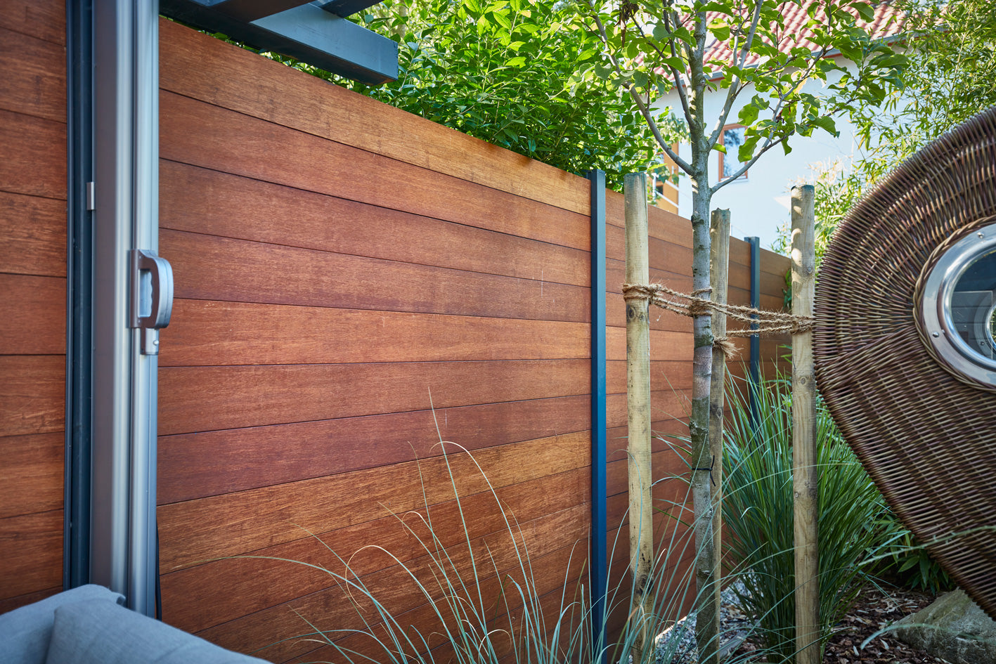 Ein moderne Bambuszaun ambooo Acapulco in warmer Holzoptik als Sichtschutz neben einer Terrasse mit Pflanzen und geflochtenem Outdoor-Möbel.