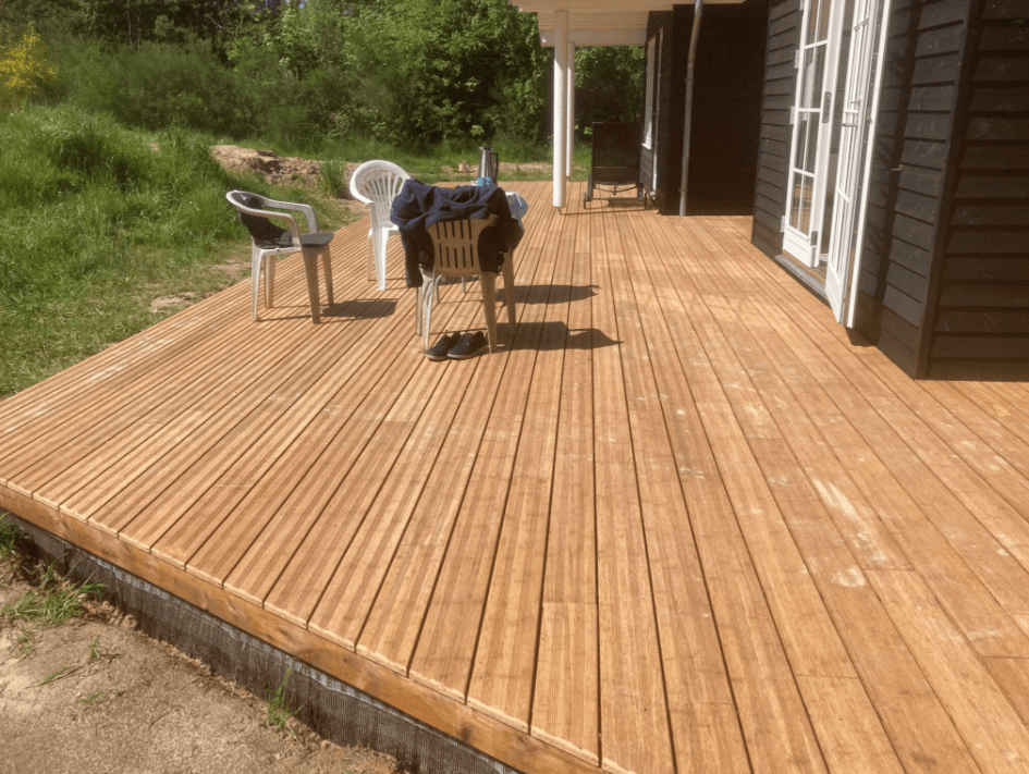 Große helle Bambusterrasse mit längs verlegten Terrassendielen vor einem Wohnhaus, möbliert mit weißen Gartenstühlen und einem Tisch, umgeben von Grün.