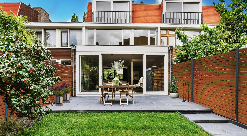 Moderner aMBOOO Bambus Sichtschutzzaun mit horizontalen Bambuslamellen in einem stilvollen Garten mit Terrasse, Rasen und moderner Hausfassade im Hintergrund.