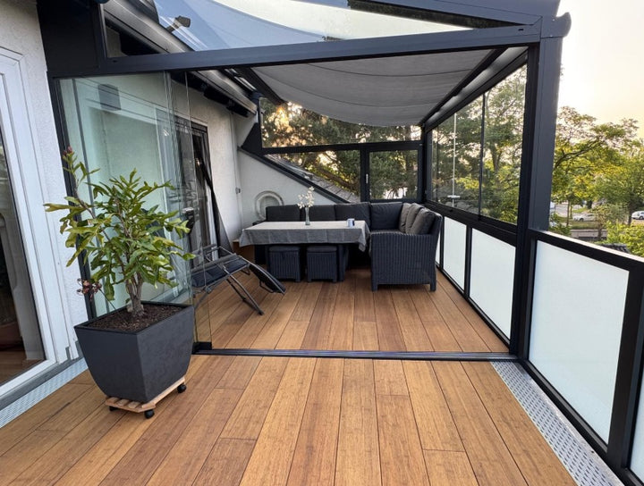 Die aMBOOO Closed Decking Bambusdiele Cristobal  unter moderner Überdachung an einem Einfamilienhaus, ausgestattet mit Gartenmöbeln. Nachhaltige Bambusdielen mit natürlicher Holzoptik, ideal für stilvolle und wetterfeste Terrassengestaltung.