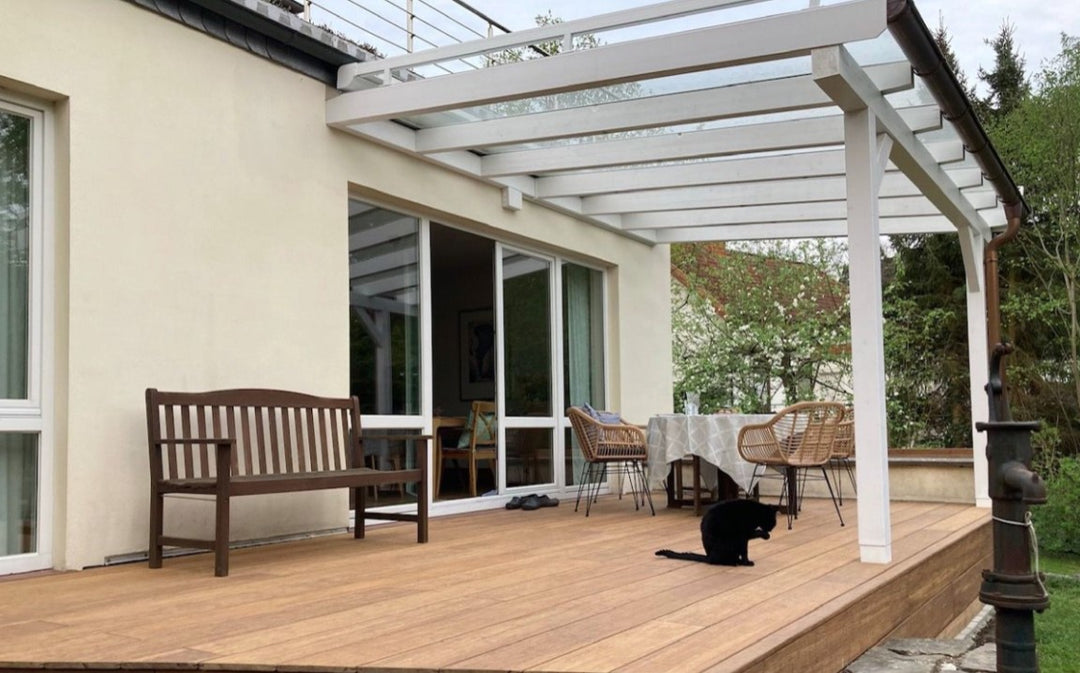 Moderne AMBOOO Bambus-Terrasse an einem Einfamilienhaus mit weißer Terrassenüberdachung aus Glas und Aluminium. Auf der großzügigen Terrasse befinden sich eine Holzbank, ein Esstisch mit Korbstühlen sowie eine schwarze Katze. Große Fensterfronten verbinden den Wohnbereich mit dem Außenbereich, während der Garten und blühende Bäume eine ruhige, wohnliche Atmosphäre schaffen.