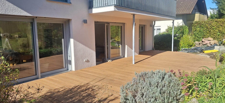 Großzügige Terrasse mit Terrassendielen aus Bambus in der Farbe Sand, an einem modernen Einfamilienhaus mit bodentiefen Fenstern, überdachtem Sitzbereich und angrenzendem Garten.
