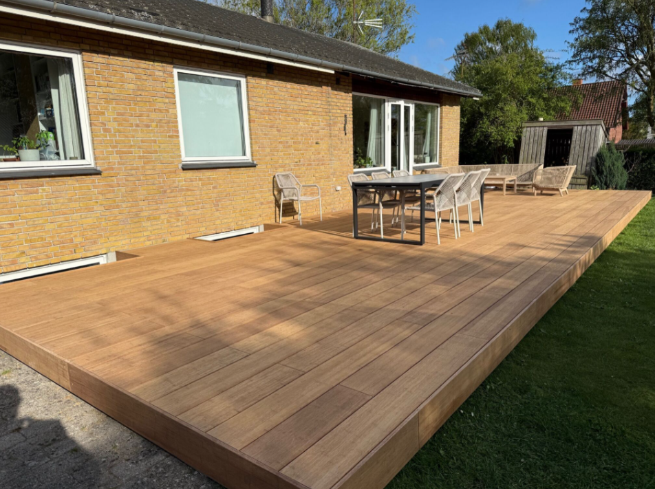 Großzügige Terrasse aus Closeddecking aMboooBambus Terrassendielen in natürlicher Holzoptik, ausgestattet mit modernem Essbereich und Lounge-Möbeln, vor einem Einfamilienhaus mit gelber Klinkerfassade – pflegeleicht und wetterbeständig 