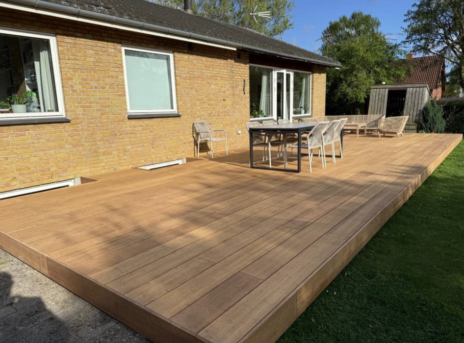 Terrasse erstellt mit ambooo closed decking Terrassendielen in natürlicher Holzoptik, aussgestattet mit moderenen Essbereich und Loungemöbeln, vor einem Einfamilienhaus mit gelber Klinkerfassade.