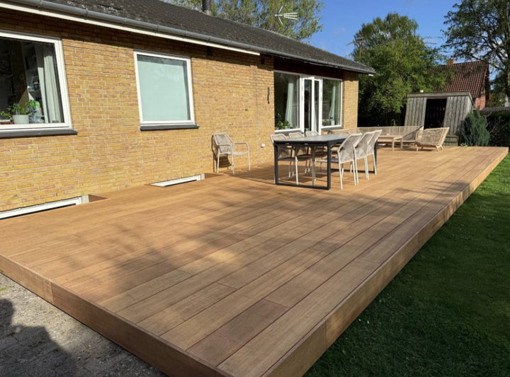 Terrasse erstellt mit ambooo closed decking Terrassendielen in natürlicher Holzoptik, aussgestattet mit moderenen Essbereich und Loungemöbeln, vor einem Einfamilienhaus mit gelber Klinkerfassade.