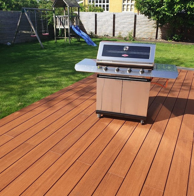 Edelstahl-Gasgrill auf einer modernen Holzterrasse in einem gepflegten Familiengarten mit Rasen, Schaukel und Rutsche bei sonnigem Wetter.