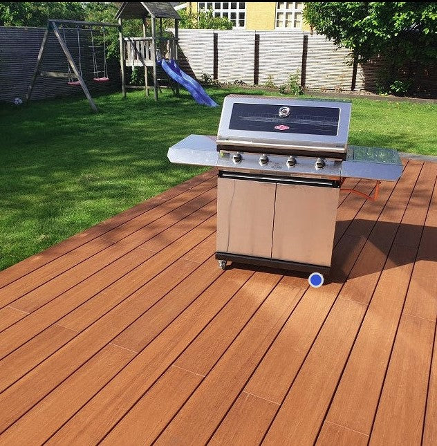 Edelstahl-Gasgrill auf einer modernen Holzterrasse in einem gepflegten Familiengarten mit Rasen, Schaukel und Rutsche bei sonnigem Wetter.
