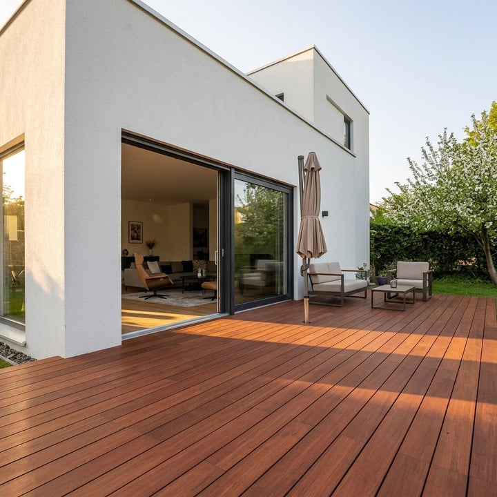 Großzügige Holzterrasse vor einem modernen weißen Wohnhaus mit Sitzgruppe, geschlossenem Sonnenschirm und geöffnetem Zugang zum Wohnbereich.