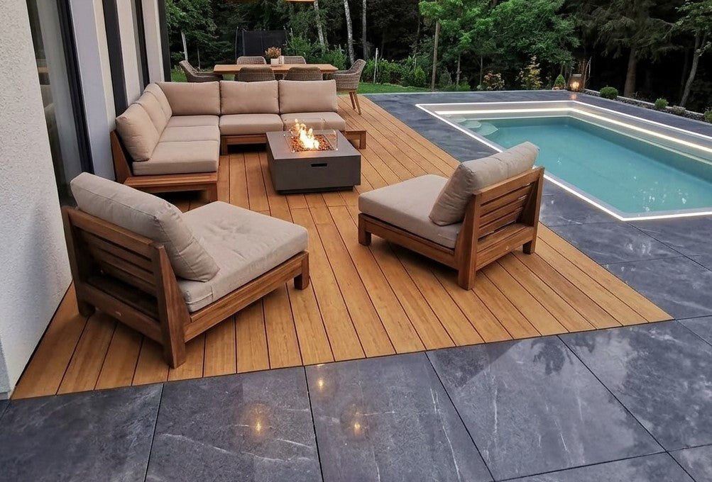 Moderne Poolterrasse mit Holzdeck, gepolsterten Loungemöbeln und brennender Feuerstelle, umgeben von dunklen Steinplatten und Wald.
