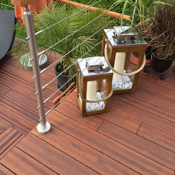 Detailaufnahme einer AMBOOO Terrasse aus rötlich-braunen Holzdielen mit moderner Edelstahlseil-Brüstung. Zwei dekorative Holzlaternen mit Kerzen und weißen Ziersteinen stehen zwischen verschiedenen Kübelpflanzen und verleihen dem Außenbereich eine stilvolle, mediterrane Atmosphäre.