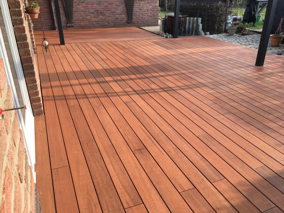Großzügige Bambusterrasse aus rötlich-braunen Terrassendielen an einer Backsteinfassade, teilweise überdacht von einer modernen Pergola mit dunklen Pfosten. Die Dielen sind längs verlegt und wirken gepflegt, während Sonnenlicht und Schattenmuster über die Fläche fallen. Im Hintergrund befinden sich weitere Backsteinwände, Pflanzgefäße und ein Gartenbereich.