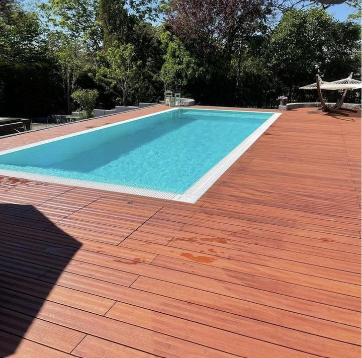 Moderner Außenpool mit großzügiger Umrandung aus rötlich-braunen Bambusdielen. Das klare, türkisfarbene Wasser bildet einen starken Kontrast zum warmen Holzdeck. Die Poolterrasse ist von üppigen Bäumen und Sträuchern umgeben, während im Hintergrund eine Hängematte zum Entspannen einlädt.