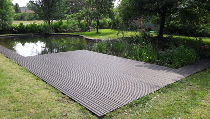 Eine weitläufige Terrasse mit schwarzen, fein geriffelten Terrassendielen erstreckt sich entlang eines naturnah gestalteten Gartenteichs. Das ruhige Wasser spiegelt die umliegenden Bäume und Sträucher wider. Rund um den Teich sorgen Rasenflächen, Schilfpflanzen und dichtes Grün für eine harmonische Gartenatmosphäre. Die Terrasse bietet einen direkten Zugang zum Wasser und fügt sich nahtlos in die natürliche Umgebung ein.