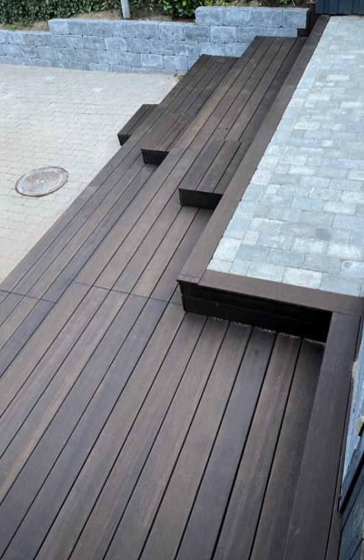 Eine moderne Terrassenanlage mit dunkelbraunen Terrassendielen in geradliniger Verlegung. In die erhöhte Terrasse sind mehrere Sitzstufen und Sitzbänke integriert, die sich harmonisch in das Design einfügen. Angrenzend befinden sich helle Pflasterflächen und eine Natursteinmauer, die den Außenbereich strukturiert und der Anlage ein hochwertiges, modernes Erscheinungsbild verleiht.