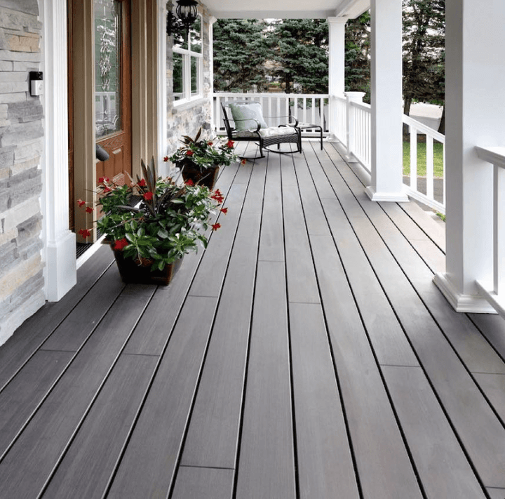 Eine überdachte Veranda mit schwarzen Ambooo Terrassendielen erstreckt sich entlang der Hausfassade. Weiße Säulen und Geländer rahmen den Außenbereich ein. Neben der Eingangstür stehen bepflanzte Blumenkübel, während am Ende der Veranda eine Sitzecke mit Sesseln zum Entspannen einlädt. Im Hintergrund sind Bäume und Gartenflächen zu sehen.